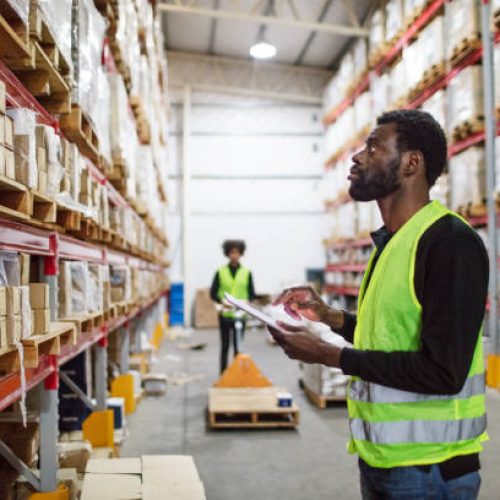 Mature warehouse worker checking inventory. African american male worker with digital tablet taking stock inventory in warehouse, with a female loader moving boxes in background.